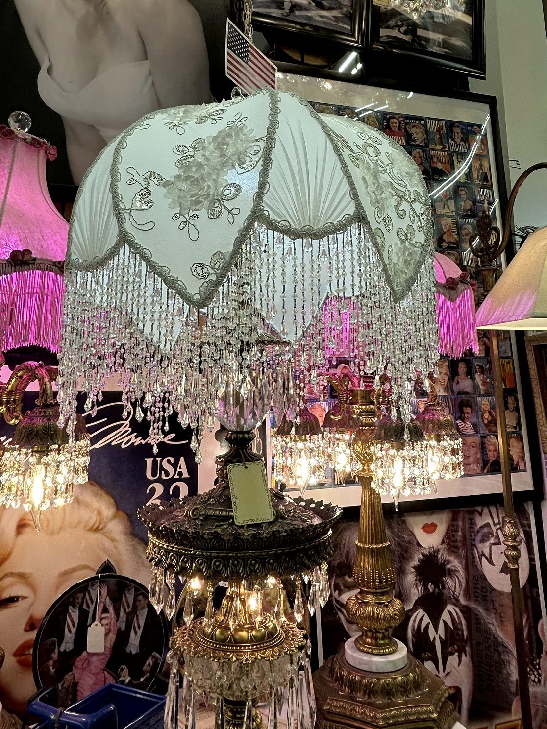 A beaded lace lampshade between two crystal chandelier lamps, with pink fringed Victorian shades glowing behind