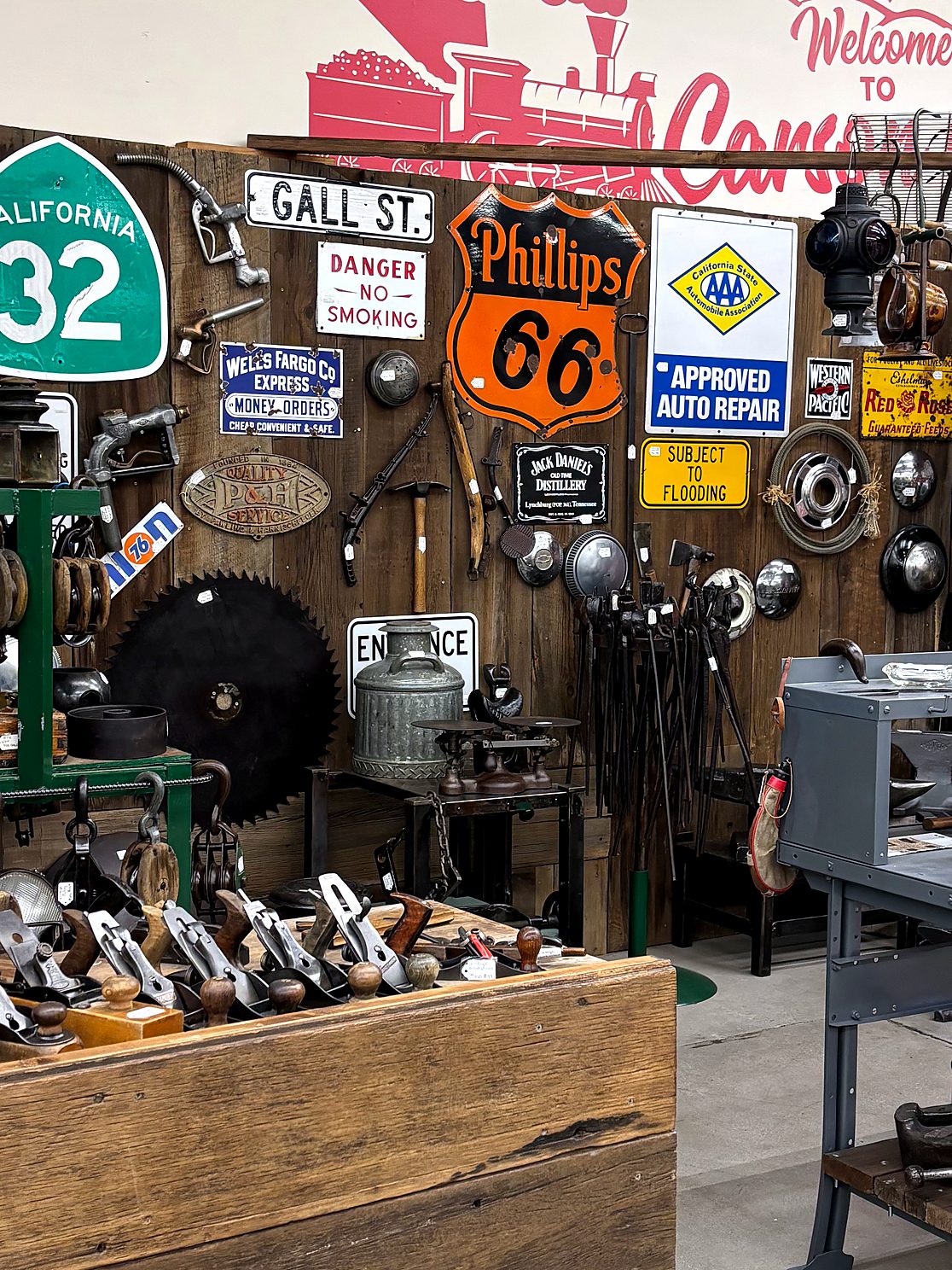 Wall of vintage signs in a vendor booth — Phillips 66, California 32, Gall St., Wells Fargo & Co. Express, AAA Approved Auto Repair, a railroad lantern, a circular saw blade, hubcaps and old hand planes