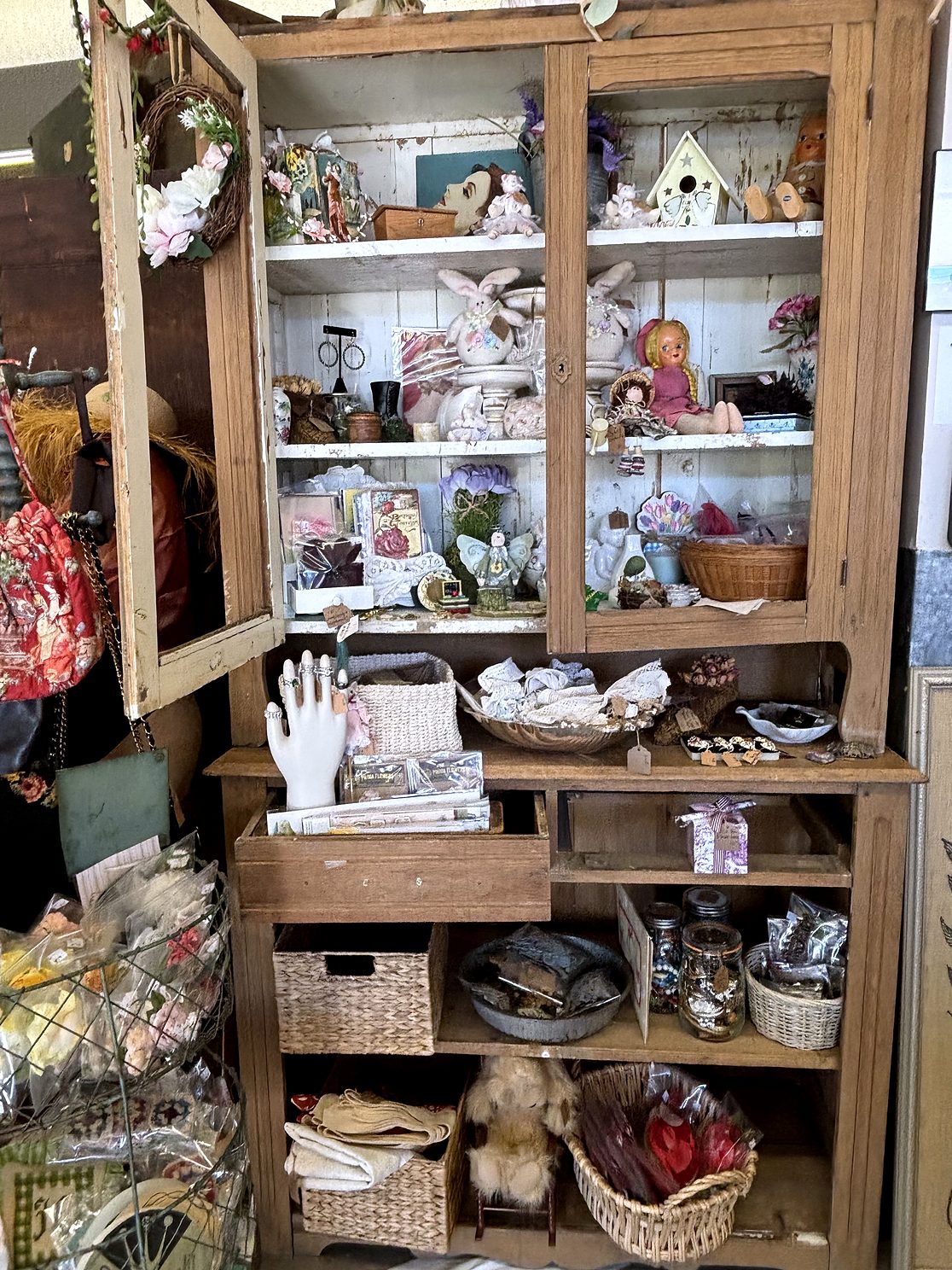 A tall wooden hutch filled with dolls, plush bunnies, a porcelain ring-holder hand, baskets and linens